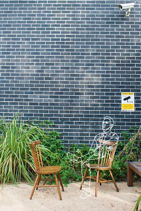 Photo of a gray-blue brick wall with CCTV camera and two chairs in a back street of Newtown, Sydney, a drawing of a man sitting in one of the chairs. By Estonian artist Anastasia Parmson